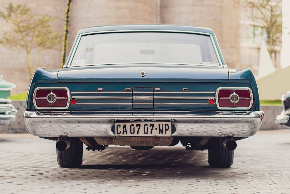 The image shows the rear view of a vintage blue Ford car parked outdoors on a paved surface, with the trunk lid, tail lights, and chrome bumper clearly visible. The vehicle features a registration plate reading 'CA 07 07 WP', and the chrome bumper exhibits some minor marks. In the background, there are blurred elements of a modern urban environment, including trees, a brick wall, and a few other parked cars, indicating a residential or city setting. The lighting suggests it is daytime with natural light illuminating the scene. This image exemplifies the careful handling and transportation of classic vehicles, relevant to house removals or furniture transport and the logistical considerations for moving valuable or vintage items, as provided by Man with Van Old Ford during home relocations and packing processes involving intricate objects like vintage cars.