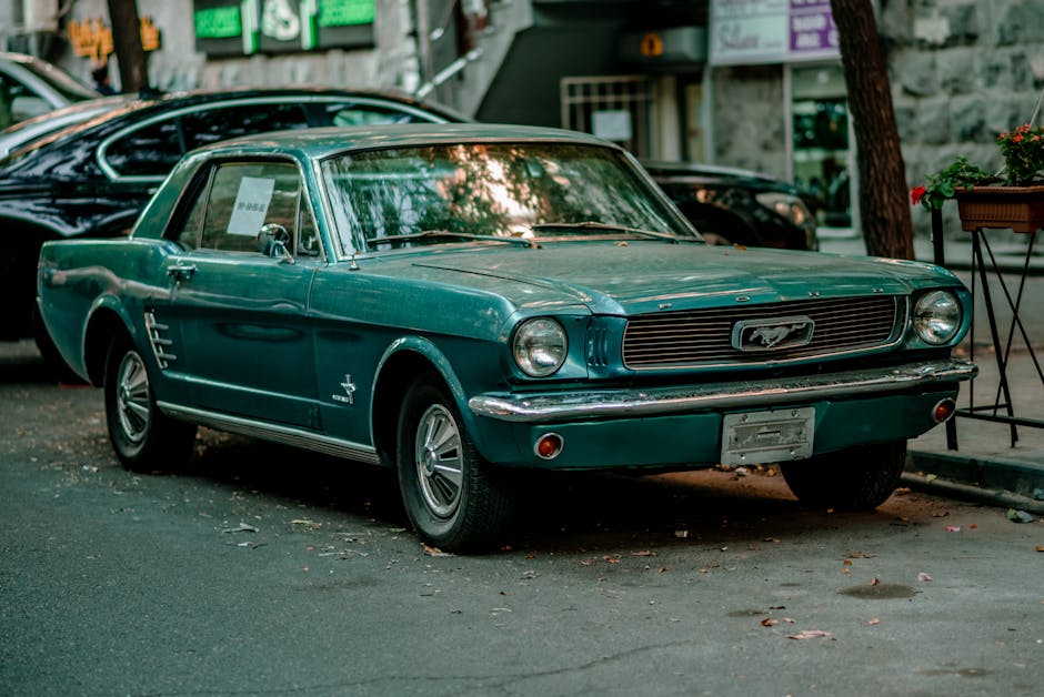 A vintage turquoise Ford Mustang parked on a city street during daylight hours, with a parked black car behind it and a shopfront with signs in the background. The car features a classic design with a prominent front grille, round headlights, and a sleek, curved body. It is positioned on the pavement next to a wooden planter box with red flowers and a utility pole, with fallen leaves scattered around on the ground. The scene suggests an urban environment where vehicle mobility and careful parking are important considerations in house removals and furniture transport. Man with Van Old Ford’s services focus on efficient packing and loading, often involving careful vehicle and furniture handling within narrow streets or old Terrace settings.