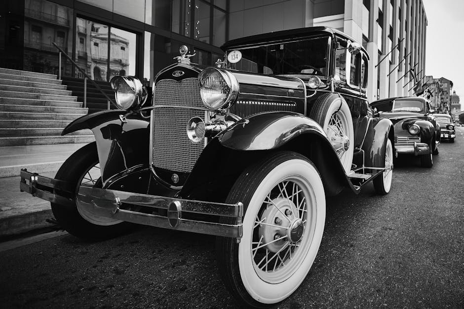 The image shows the rear view of a vintage blue Ford car parked outdoors on a paved surface, with the trunk lid, tail lights, and chrome bumper clearly visible. The vehicle features a registration plate reading 'CA 07 07 WP', and the chrome bumper exhibits some minor marks. In the background, there are blurred elements of a modern urban environment, including trees, a brick wall, and a few other parked cars, indicating a residential or city setting. The lighting suggests it is daytime with natural light illuminating the scene. This image exemplifies the careful handling and transportation of classic vehicles, relevant to house removals or furniture transport and the logistical considerations for moving valuable or vintage items, as provided by Man with Van Old Ford during home relocations and packing processes involving intricate objects like vintage cars.