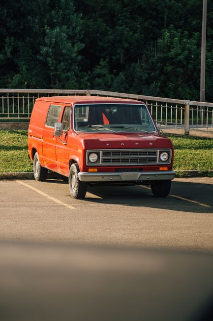 A red Ford commercial van with a boxy design, parked in an outdoor parking lot marked with yellow lines. The van has grey side mirrors, a front grille with horizontal slats, and rectangular headlights