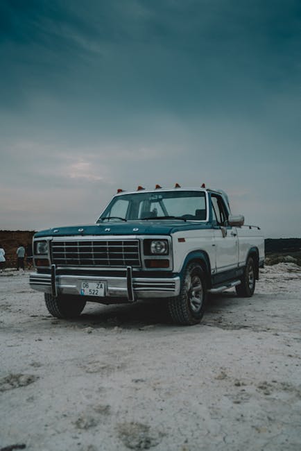 A vintage Ford pickup truck with a white body and dark blue roof, parked on a barren, dusty area under a cloudy sky. The truck has a sturdy front grille, round headlights, and orange warning lights on top of the cab. Inside the truck bed, there are no visible items. The surrounding environment appears to be an open, desolate landscape with no buildings or other vehicles nearby. This image captures a classic vehicle that could be used in home relocation or furniture transport services, similar to those provided by Man with Van Old Ford. The scene emphasizes the durability and reliability of vehicles used in moving and removal operations, set against a natural, outdoor backdrop.
