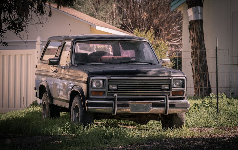 A weathered, black vintage van parked on a grassy area in a suburban backyard, with a white fence, a tree with a metal pole nearby, and a small building with a sloped roof in the background. The van’s front grille and bumper are visible, with signs of rust and dirt on its bodywork. The vehicle appears to be in a stationary position, possibly awaiting removal or transportation as part of house clearance or furniture transport during a home relocation. The scene is lit with natural daylight, highlighting the aged condition of the van and surrounding environment, which includes some grass and uneven ground. This image reflects the logistics involved in furniture transport or bulk waste removal services provided by companies like Man with Van Old Ford during the moving process, especially when handling bulky items or old vehicles as part of an overall move or clearance project.