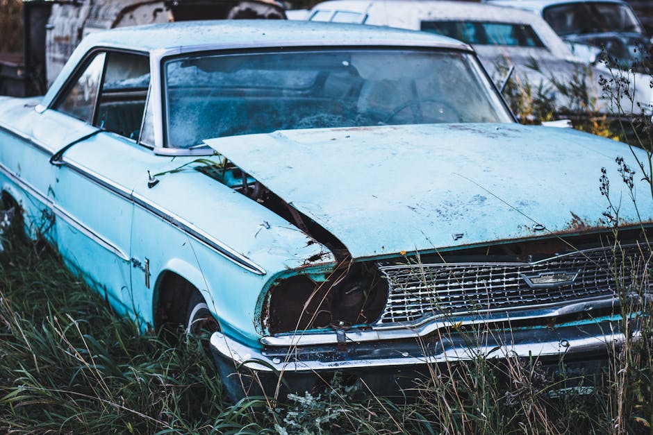 A weathered, light blue vintage car with significant body damage and missing headlights, situated in tall grass and weeds on a patch of outdoor land. The car's hood is partially open, revealing internal components, and the metal surfaces are rusted and covered with dirt. The windows are intact but appear dirty, and the vehicle is surrounded by overgrown vegetation. In the background, there are several other cars parked closely together in what appears to be an outdoor storage or scrap yard area. The setting suggests an abandoned or decommissioned vehicle awaiting disposal or removal. For house removals or furniture transport, Man with Van Old Ford handles logistical aspects of clearing and disposing of old vehicles or bulky items during property clearance or relocation processes.