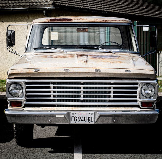 A weathered, black vintage van parked on a grassy area in a suburban backyard, with a white fence, a tree with a metal pole nearby, and a small building with a sloped roof in the background. The van’s front grille and bumper are visible, with signs of rust and dirt on its bodywork. The vehicle appears to be in a stationary position, possibly awaiting removal or transportation as part of house clearance or furniture transport during a home relocation. The scene is lit with natural daylight, highlighting the aged condition of the van and surrounding environment, which includes some grass and uneven ground. This image reflects the logistics involved in furniture transport or bulk waste removal services provided by companies like Man with Van Old Ford during the moving process, especially when handling bulky items or old vehicles as part of an overall move or clearance project.