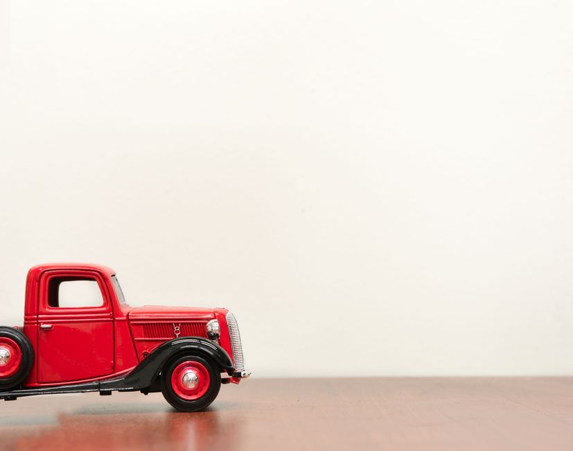 A detailed close-up of a vintage red toy truck with black fenders and red wheels sits on a wooden surface against a plain white background. The truck's design includes a rounded cabin, silver grille, 
