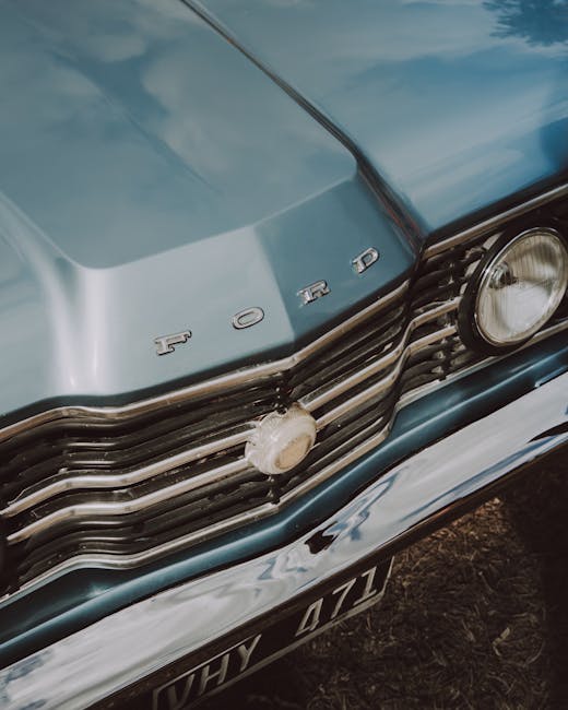 Close-up view of the front grille and hood of a vintage Ford vehicle, featuring chrome accents and a central emblem with the word 'FORD' on the hood. The headlight is visible on the right side, with p