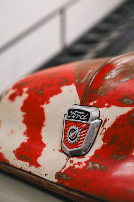 Close-up view of the front grille and hood of a vintage Ford vehicle, featuring chrome accents and a central emblem with the word 'FORD' on the hood. The headlight is visible on the right side, with p