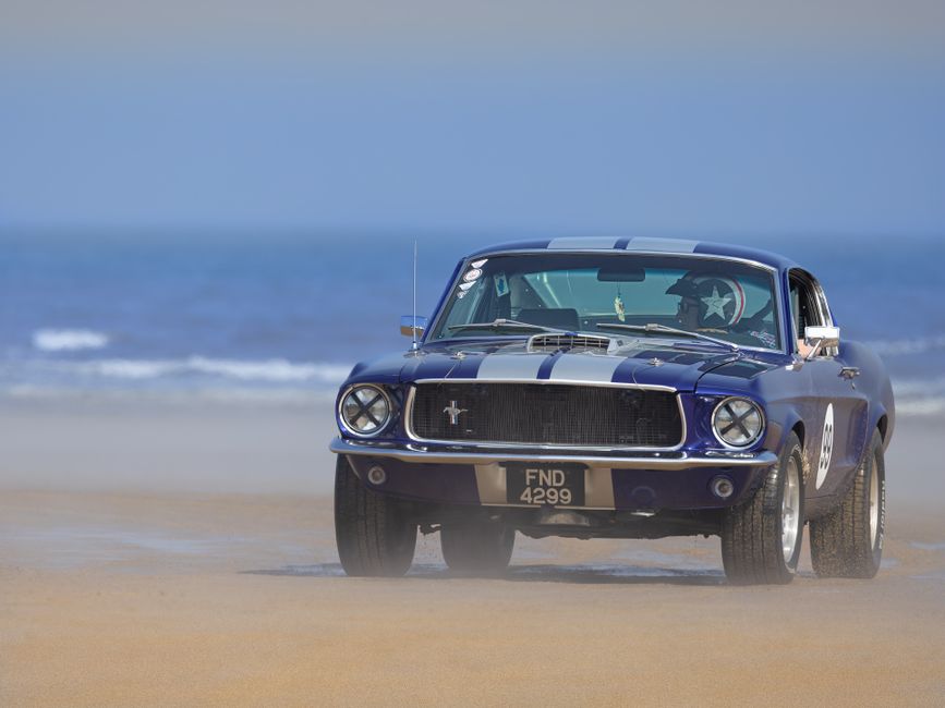 A blue vintage Ford Mustang with racing stripes parked on a sandy beach near the shoreline, with calm waves and a clear sky in the background. The car features a classic grille with the Mustang emblem, round headlights, and a black front grille. The vehicle's tires are slightly dusted with sand, indicating recent movement on the beach surface. The windshield displays a star-shaped window decal, and there are objects like a small hanging ornament inside the car visible through the glass. The scene shows the car positioned close to the water, suggesting a leisure drive or a scenic stop during a home relocation journey that involves transporting vehicles as part of a house move. Man with Van Old Ford, a professional removals service, may handle such transport logistics for clients moving houses or requiring vehicle transportation during their move.