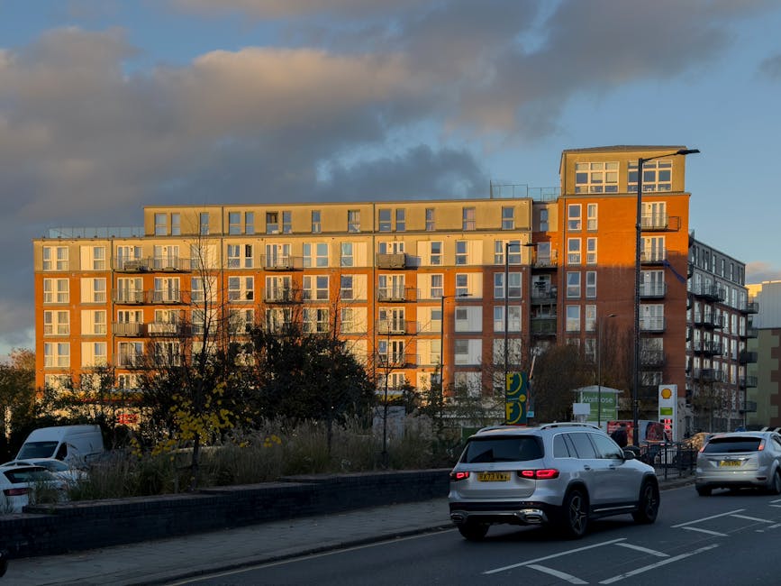 A multi-storey apartment building with a modern design, featuring numerous large windows and small balconies, is illuminated by the warm glow of late afternoon or early evening sunlight. The building's exterior showcases a combination of orange and beige in a contemporary architectural style. In the foreground, a street with moving traffic includes several vehicles, such as a white van and a grey SUV, traveling along marked lanes. To the left, a sidewalk bordered by trees and low shrubs separates the roadway from a parking area, where a few parked cars are visible. Nearby, a petrol station with green and yellow signage is situated close to the building. The scene captures a typical urban environment involved in home relocation and furniture transport activities, illustrating the logistics of moving services as part of house removals at Old Ford. The lighting and shadows emphasize the textures of the building materials and the street scene, aligning with the context of packing, loading, and transportation processes facilitated by Man with Van Old Ford.