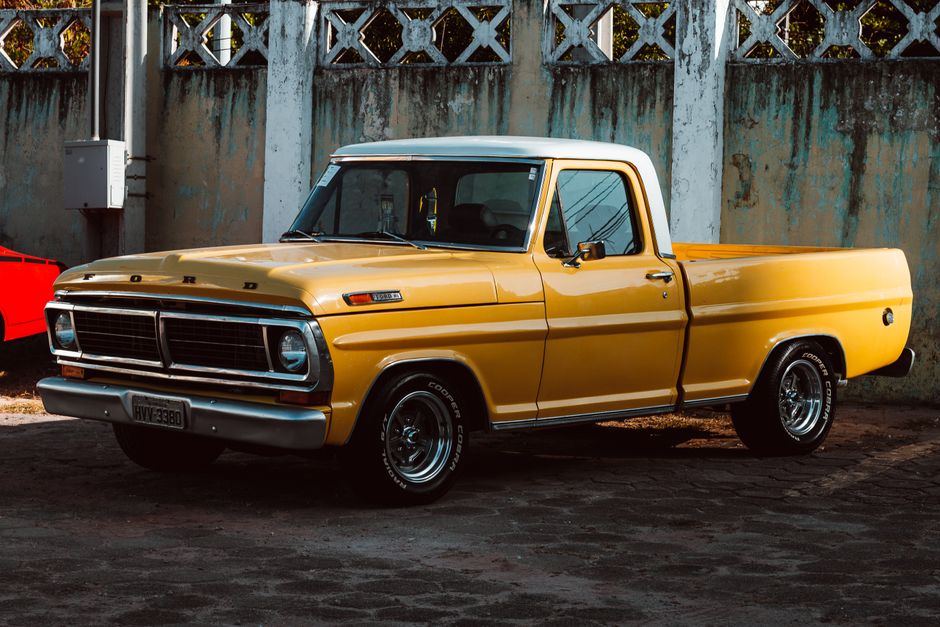 A dilapidated, light blue vintage car with visible rust and damage, parked amidst tall grass and weeds, surrounded by other abandoned vehicles in the background. The front grille and headlights are missing or broken, with the hood slightly ajar. The car appears to be unused and left outdoors in a neglected state, situated near a driveway or parking area. This setting reflects an environment where salvage or removal services, like those offered by Man with Van Old Ford, may be necessary for home relocation or vehicle collection processes involving old or damaged vehicles.