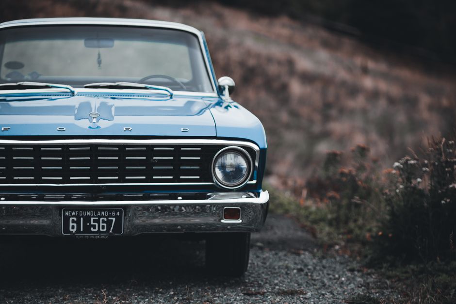Close-up view of the front of a classic blue Ford car parked on a gravel driveway. The vehicle features a black grille, round headlights, and a visible front license plate from Newfoundland with the r