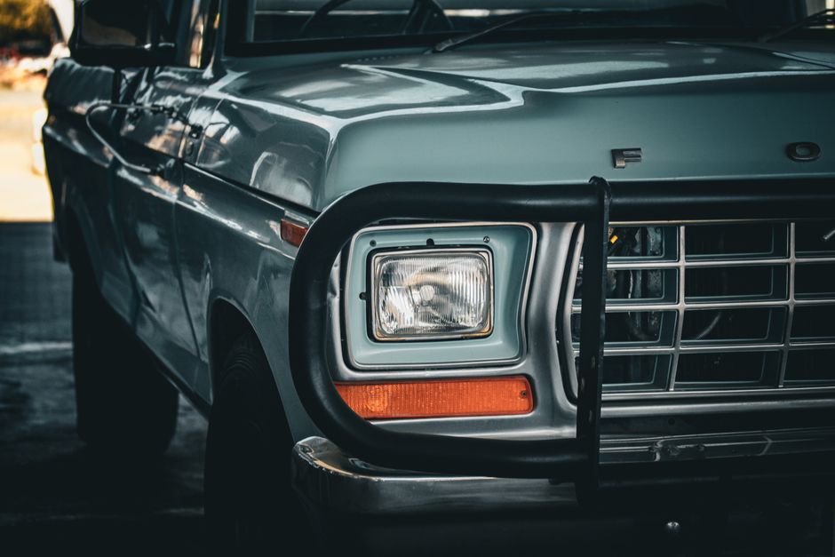 Close-up view of the front of a classic blue Ford car parked on a gravel driveway. The vehicle features a black grille, round headlights, and a visible front license plate from Newfoundland with the r