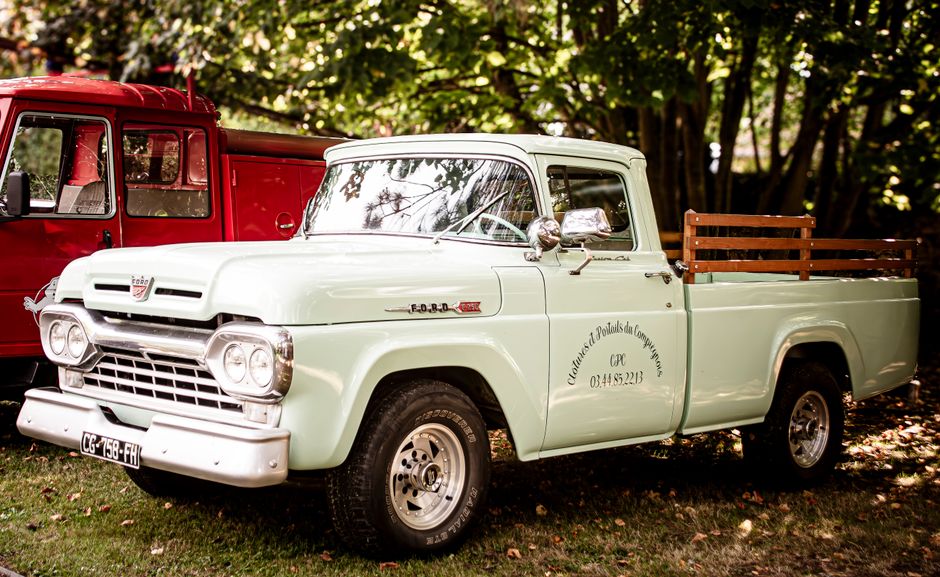 A vintage light mint green pickup truck parked outdoors on a grassy area with fallen leaves, surrounded by trees with green foliage. The truck features a wooden side rail on the bed, a chrome side mirror, and a classic front grille with dual headlights. The vehicle displays black text on the door indicating it is associated with a company involved in house removals and furniture transport, with the number 0344 852213. Behind the mint green truck, there is a partially visible red truck, suggesting a relocation or transportation scenario. The background of dense trees provides natural lighting, with sunlight filtering through the leaves, and the scene captures the loading or unloading process in a residential or park setting, consistent with the context of home relocation services provided by [COMPANY_NAME].
