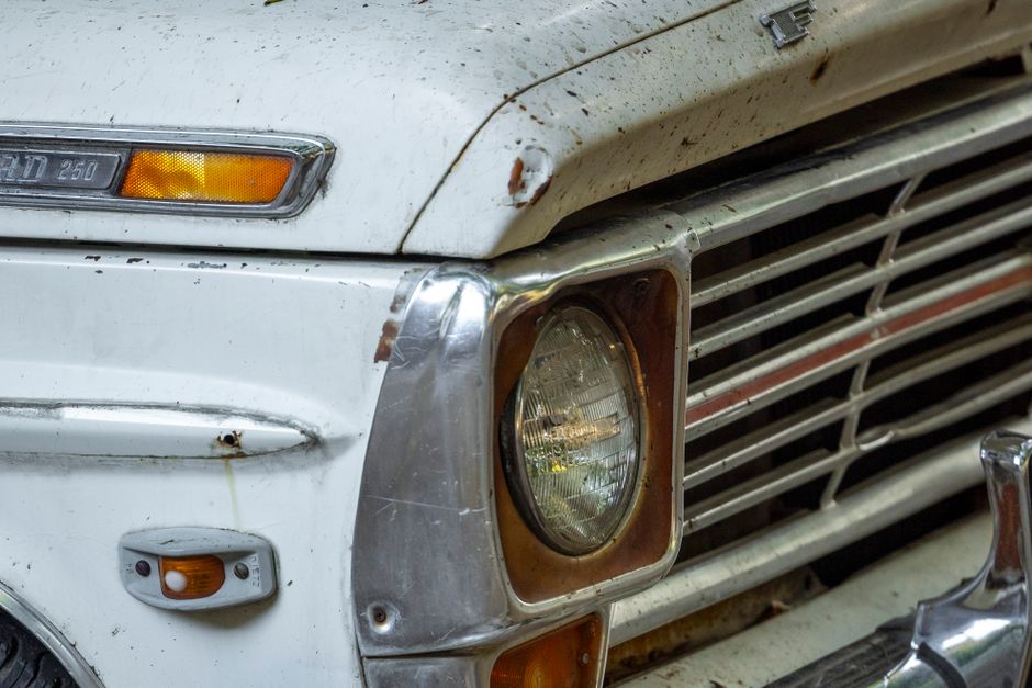 A vintage white Ford pickup truck with a black grille and bumper, parked on a rough, sandy surface under a partly cloudy sky. The truck features a double cab design with four doors, black side mirrors, and a small side window behind the front doors. The vehicle has a roof-mounted light bar with orange warning beacons. In the background, a few people are visible near the horizon, and the landscape appears arid and open. The scene is well-lit with natural daylight, and the truck is positioned facing slightly to the left, illustrating its interior cabin and the flatbed cargo area. This image reflects aspects of vehicle transport and logistics relevant to home relocation or moving services offered by companies such as Man with Van Old Ford.