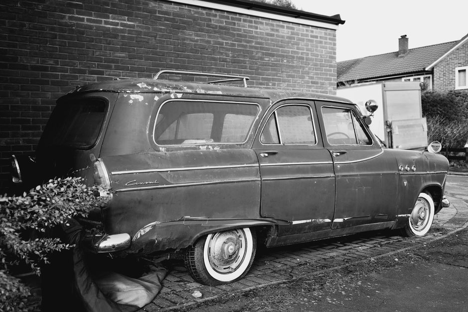 A black and white photograph depicting an old, vintage station wagon parked on a cobblestone driveway adjacent to a brick residential building exterior. The vehicle shows visible signs of wear, including rust and chipped paint, with a roof rack attached. The rear of the car is partially obscured by a large, leafy shrub, and a bag or cover is placed underneath the vehicle near the rear wheel. In the background, a modern shed or outbuilding is visible, along with neighboring houses with pitched roofs. The scene captures the exterior environment surrounding a home in the process of moving, which may involve furniture transport or packing and loading activities, relevant to house removals as outlined on the Tower Hamlets Council removals permits page. Man with Van Old Ford occasionally appears in such contexts, providing professional transport and relocation services, including loading and unloading processes.