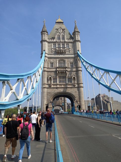 The image shows a group of people standing on the sidewalk of Tower Bridge in London, with the iconic Victorian Gothic style tower featuring detailed stonework, turrets, and decorative elements, flanked by the blue and white suspension cables of the bridge. The pedestrians include men and women of various ages, some carrying backpacks and wearing casual clothing, standing near the base of the bridge’s central tower. The scene is set during daylight with clear weather, and the surrounding cityscape is visible in the background. This view exemplifies the urban environment where home relocation or furniture transport services such as those provided by Man with Van Old Ford might be required for residents or businesses in the E3, Old Ford area, particularly when navigating or planning moves around prominent landmarks like Tower Bridge.