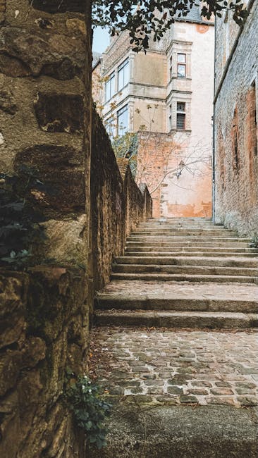 A narrow outdoor staircase with uneven stone steps ascending between a weathered stone wall on the left and a brick building on the right, leading upwards towards a courtyard or street in an older residential area. The staircase is bordered by a low stone wall, with some greenery and small plants growing at its base. In the background, multi-storey buildings with classic architectural features are visible, including windows and balconies, under a partly cloudy sky. The scene, seen from the bottom of the stairs, suggests a typical setting for house removals or home relocation services, with careful navigation of tight spaces as handled by companies like Man with Van Old Ford. The natural light indicates daytime, and the weather-worn surfaces highlight the outdoor environment characteristic of historic districts.