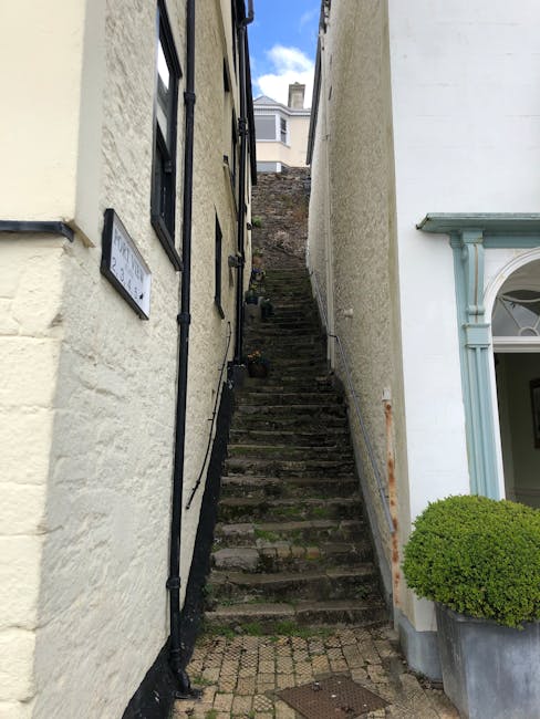 A narrow outdoor staircase made of weathered stone steps ascending between two buildings, one with a textured beige exterior wall and the other with a smooth white finish and decorative blue trim around a large window. The steps are uneven and appear aged, with some moss or dirt on their surfaces. A black downspout runs vertically along the beige wall, and a rusted metal handrail is attached to the white building on the right side. The staircase leads up to a small, partially visible area at the top, with an outdoor setting visible in the background. The ground at the base of the stairs is paved with cobblestones, and a round, well-maintained green shrub in a large concrete planter is situated to the right of the stairs, near the white building. This scene is part of a typical residential area, suitable for house removals, with clear indications of a property’s entrance and surrounding features, supported by Man with Van Old Ford's house move logistics specifications.