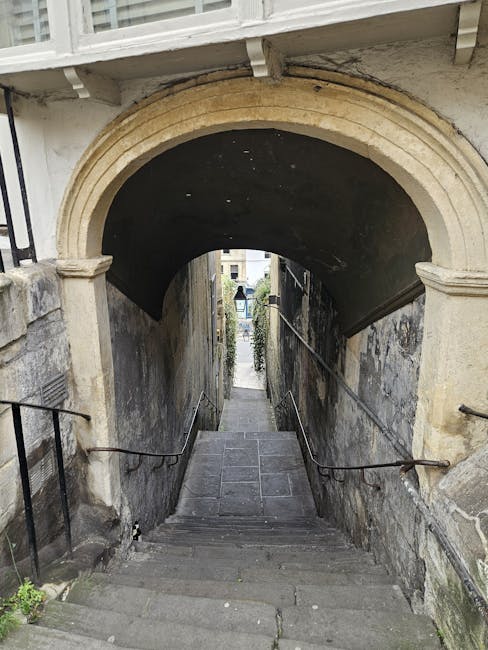 A narrow outdoor staircase with uneven stone steps ascending between a weathered stone wall on the left and a brick building on the right, leading upwards towards a courtyard or street in an older residential area. The staircase is bordered by a low stone wall, with some greenery and small plants growing at its base. In the background, multi-storey buildings with classic architectural features are visible, including windows and balconies, under a partly cloudy sky. The scene, seen from the bottom of the stairs, suggests a typical setting for house removals or home relocation services, with careful navigation of tight spaces as handled by companies like Man with Van Old Ford. The natural light indicates daytime, and the weather-worn surfaces highlight the outdoor environment characteristic of historic districts.