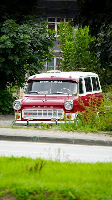 A vintage red and white Ford van parked on a residential street, surrounded by green foliage and trees. The van's front grille, headlights, and windshield are visible, with the vehicle partially obscured by nearby bushes. Behind the van, there is a house with a large window and a white frame, set among lush greenery. The pavement and grass verge are in the foreground, with the van positioned close to the curb. This scene captures a typical setting for home relocation or furniture transport, with [COMPANY_NAME] involved in the logistics of moving items safely. The lighting is natural, likely during daytime, highlighting the details of the vehicle and surrounding environment, suitable for illustrating house removals, packing, and loading processes in Old Ford as referenced on the page about moving routes and van access.