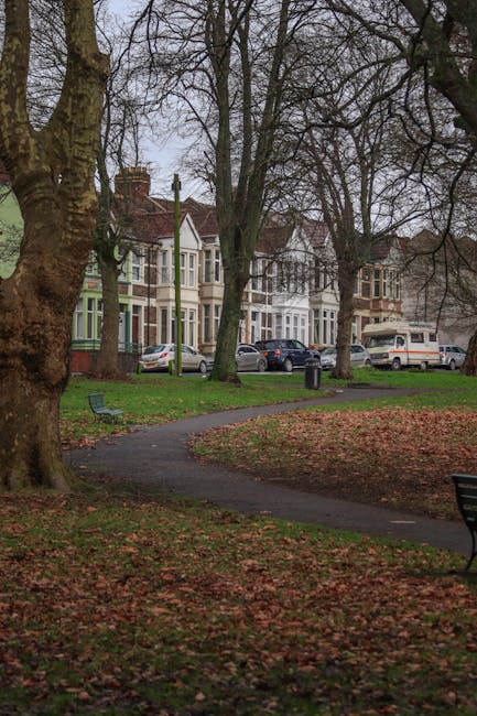 A residential street in Old Ford lined with Victorian-style terraced houses featuring bay windows and decorative brickwork, set behind a park with winding paved paths and mature leafless trees, indicating late autumn or early winter. The park has a grassy area covered with fallen leaves, a few park benches, and a small trash bin near the walking path. Visible in the background are several parked cars along the pavement, including a white van, which is partially visible through the trees. The scene shows diffuse natural lighting on an overcast day. The image relates to home relocation or furniture transport services, providing context for local house removals in Old Ford, as serviced by Man with Van Old Ford, who handles packing, moving, and logistical support in such environments.